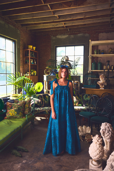 Woman in a blue dress standing in a rustic room with large windows and shelves.