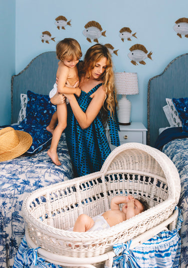 Woman in a blue patterned dress holding a child next to a baby in a white wicker crib with blue walls and fish decals.