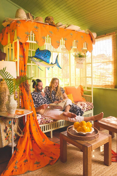 Man and woman in matching black and white palm tree patterned outfits sitting on a patterned day bed reading a magazine. 