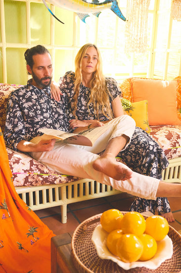 A man and a woman in matching black and white palm tree patterned outfits sitting on a patterned day bed in a colorful living room.