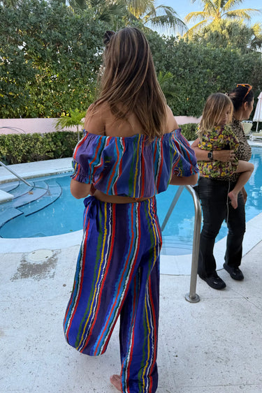 Woman in a colorful striped cropped top and bubble pants standing by a pool with two other people in the background.