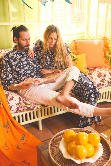 Man and woman in matching black and white palm tree patterned outfits sitting on a patterned day bed reading a magazine.