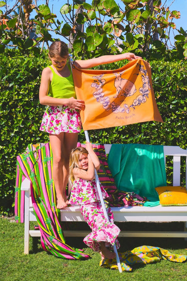 Two children wearing colorful matching floral outfits playing with a Coniglio Palm Beach flag in front of a white bench outdoors.