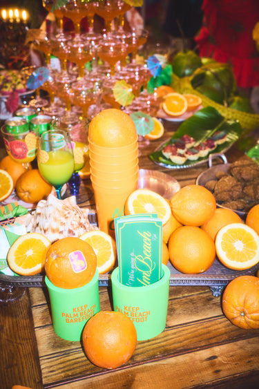 Oranges and bumper stickers  in Keep Palm Beach Barefoot koozies on a wooden table with a festive background.