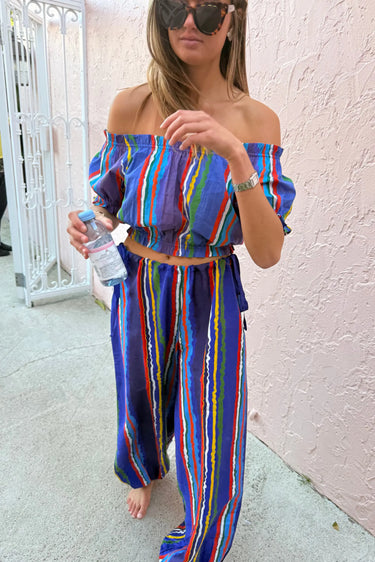 Woman wearing a colorful striped cropped top and bubble pants holding a water bottle against a light-colored wall.