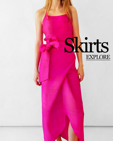 A woman modeling a bright hot pink wrap skirt with a large bow detail at the waist, set against a plain white studio background. The text 'Skirts EXPLORE' is overlaid on the right side. Links to the skirts collection.