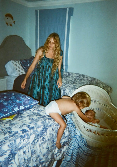 Woman in a blue patterned dress standing next to a child in a crib in a bedroom with floral bedding.
