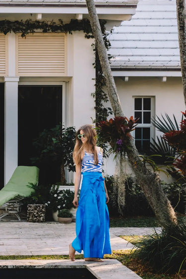 Woman in blue and white patterned top and vibrant blue wrap pants standing in front of a house with green chairs.