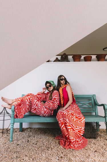 Two women in red and white patterned skirts and tops sitting on a green bench.