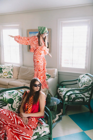 Two women in colorful red and white patterned skirts and tops standing in a room with patterned furniture and decor.