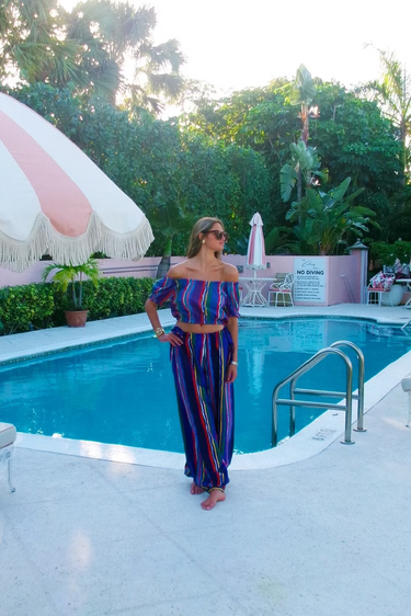 Woman in cropped top and bubble pants standing by a pool with a pink and white umbrella and greenery in the background