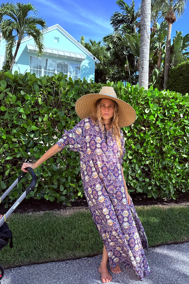 A woman wearing a blue and cream printed button down maxi dress, a wide-brimmed straw hat, and sunglasses stands barefoot outdoors in front of a green hedge and a light blue house.