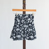 Black mini skirt with white floral and palm tree pattern hanging on a wooden hanger against a white background