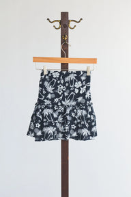 Black mini skirt with white floral and palm tree pattern hanging on a wooden hanger against a white background