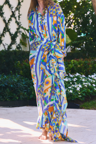 Woman wearing a colorful geometric button down and ruffle power pareo standing outdoors with greenery in the background.