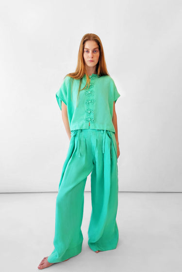 A woman modeling a mint green two-piece outfit consisting of a short-sleeve top with decorative frog closures down the front and matching wide-leg pants, set against a plain white studio background.