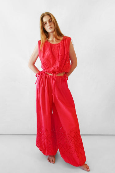 A woman modeling a bright red sleeveless top with wide-leg bubble-style pants and eyelet embroidery, standing against a plain white studio background.