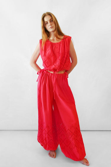 A woman modeling a bright red sleeveless top with wide-leg bubble-style pants and eyelet embroidery, standing against a plain white studio background.