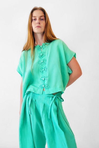 A woman modeling a bright mint green two-piece outfit consisting of a short-sleeve button-front top and matching wide-leg pants, photographed against a plain white studio background.