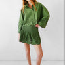 A woman modeling a matching green two-piece set consisting of a long-sleeve button-up shirt and a ruffled mini skirt, both featuring eyelet embroidery, set against a plain light gray studio background.