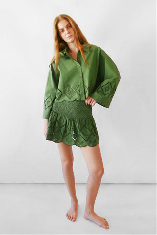 A woman modeling a matching green two-piece set consisting of a long-sleeve button-up shirt and a ruffled mini skirt, both featuring eyelet embroidery, set against a plain light gray studio background.