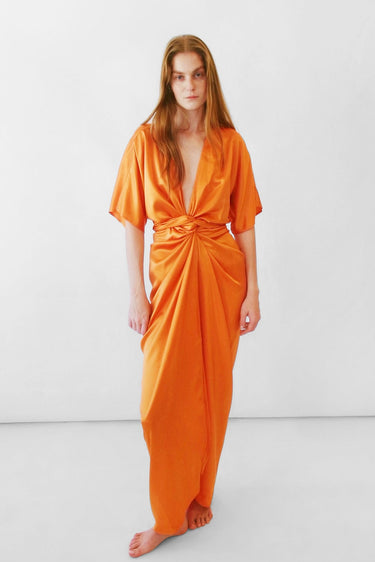 A woman modeling a bright orange silk charmeuse maxi dress with short flutter sleeves, a deep V-neckline, and a gathered twist detail at the waist, standing against a plain white studio background.