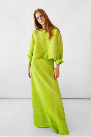 A model with long red hair wearing a bright lime green silk two-piece set consisting of a cropped long-sleeve top and a matching maxi skirt, set against a plain light gray studio background.