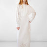 A woman modeling a long white dress with eyelet embroidery on the sleeves and hem, standing against a plain white studio background.