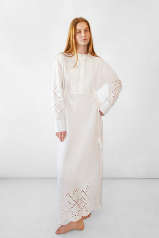 A woman modeling a long white dress with eyelet embroidery on the sleeves and hem, standing against a plain white studio background.