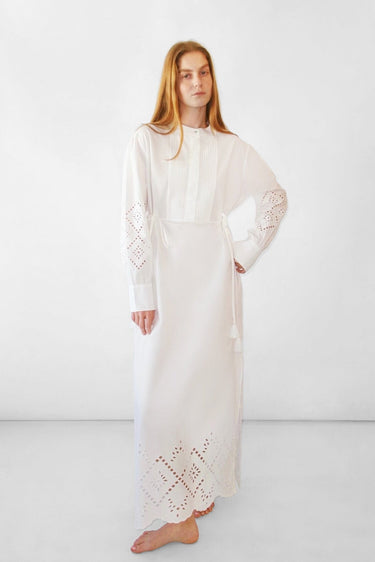 A woman modeling a long white dress with eyelet embroidery on the sleeves and hem, standing against a plain white studio background.