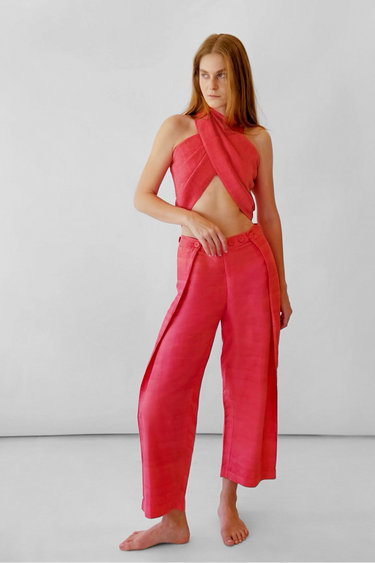A model stands barefoot in a studio wearing a bright coral pink two-piece outfit consisting of a halter wrap-style crop top and matching wide-leg pants.