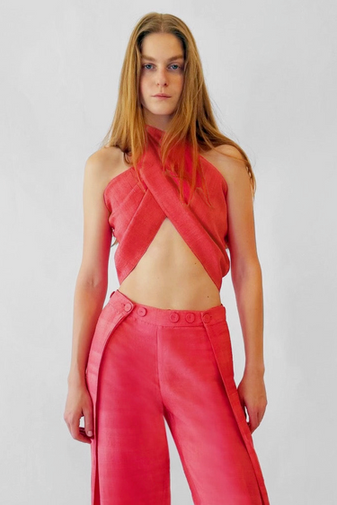 A model wearing a vibrant coral pink two-piece outfit consisting of a halter crop top and matching high-waisted buttoned trousers, set against a plain light gray background.