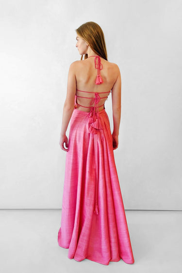 A woman stands with her back to the camera wearing a bright pink sleeveless top with a strappy open back and tassel details and a long skirt, photographed in a studio setting.