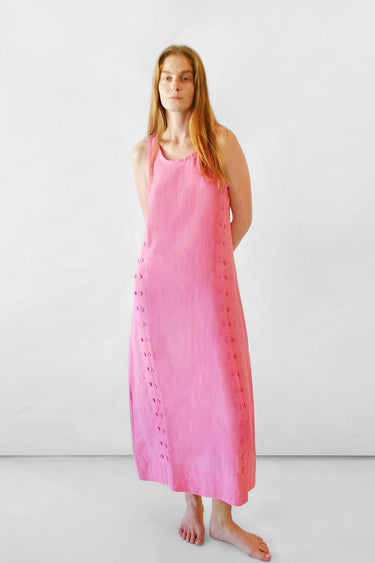 A woman stands barefoot against a plain white studio background wearing a sleeveless pink midi dress in linen.